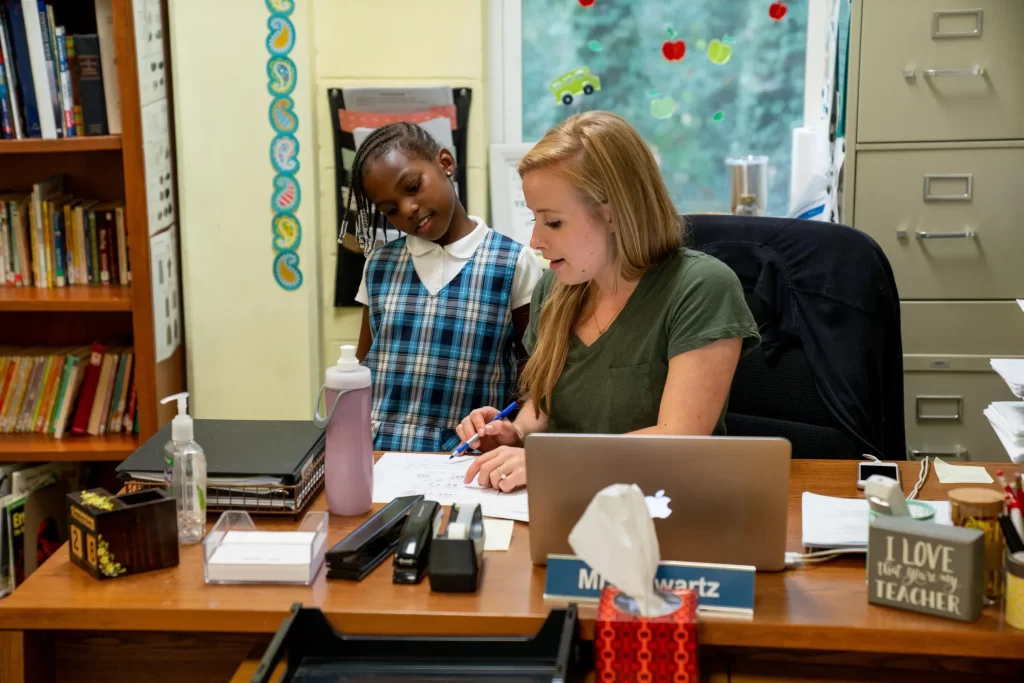 student standing at a teacher's desk while she explains a concept